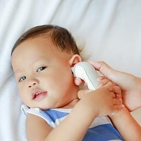 Mother takes temperature for baby boy with ear thermometer on bed at home.